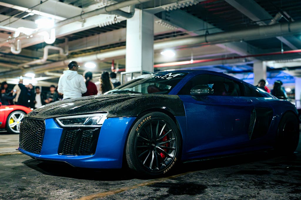 Imagen actual: Sleek blue supercar with carbon fiber detailing at a car meet in a dimly lit garage.