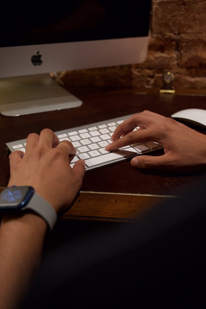 Imagen actual: Hands typing on a desktop keyboard with a smartwatch visible, indoors.